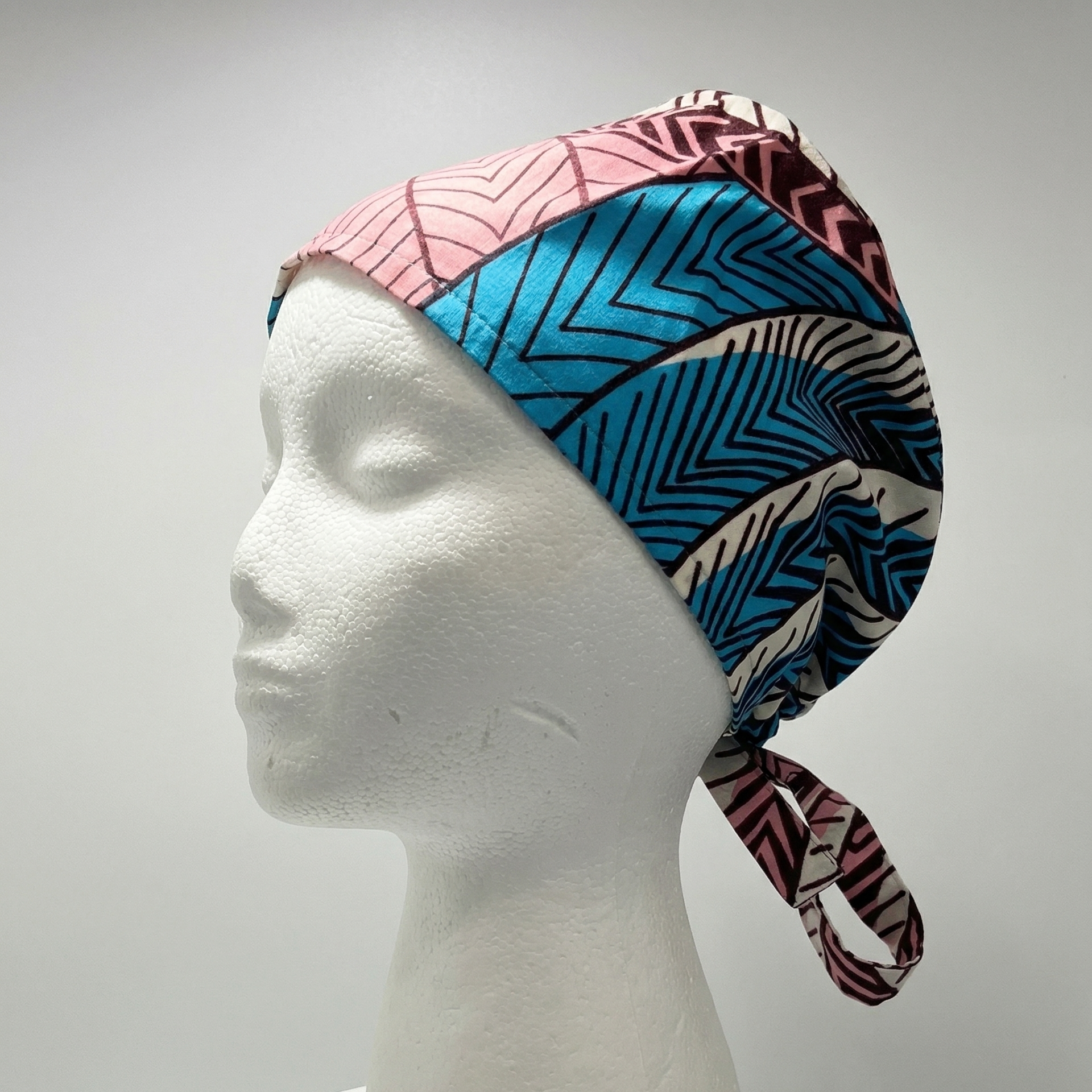 A close-up studio photograph of a manikin head displaying a patterned scrub cap. The cap features a geometric print with various sections, including a pink area with fine lines, a dominant blue-green chevron pattern, and other dark and gold geometric shapes. The tail of the cap is visible, showing a purple-maroon abstract pattern.