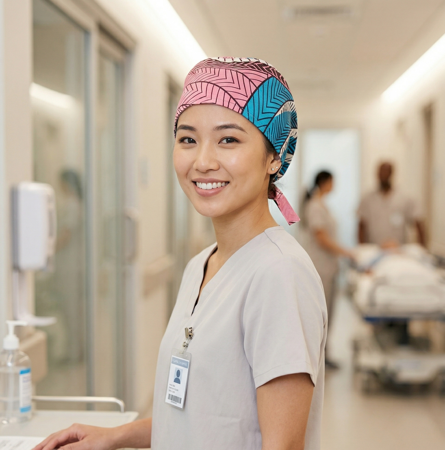 A photograph of the same woman from the second image, wearing the same scrubs and patterned scrub cap, now posed in a hospital corridor. She is looking and smiling towards the camera, while other blurry figures (a doctor and medical staff moving a patient) are visible in the background, creating context for her role in a medical environment.