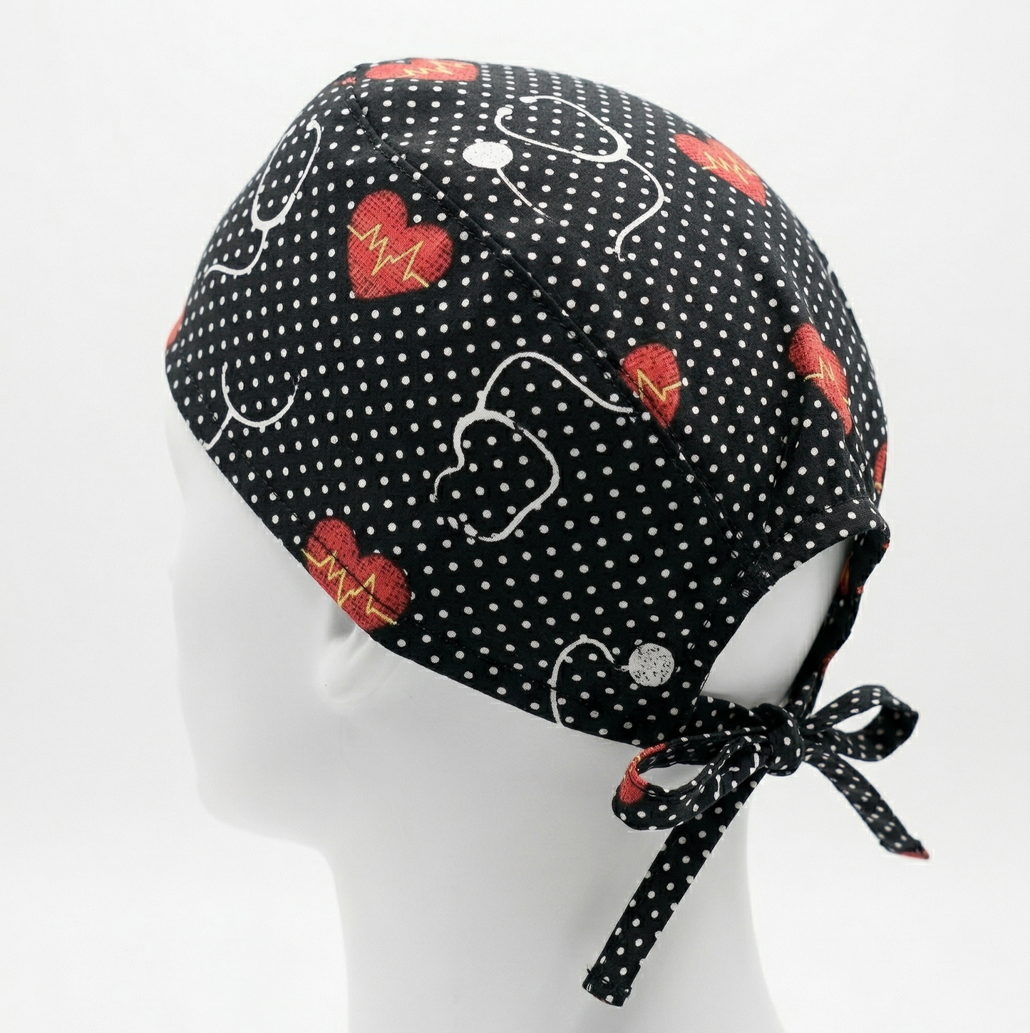 Black Stethoscope Surgical Cap 1: A studio photograph featuring a black surgical cap with a pattern of white stethoscopes and red hearts, displayed on a white mannequin head against a light grey background. The back ties of the cap are clearly visible.