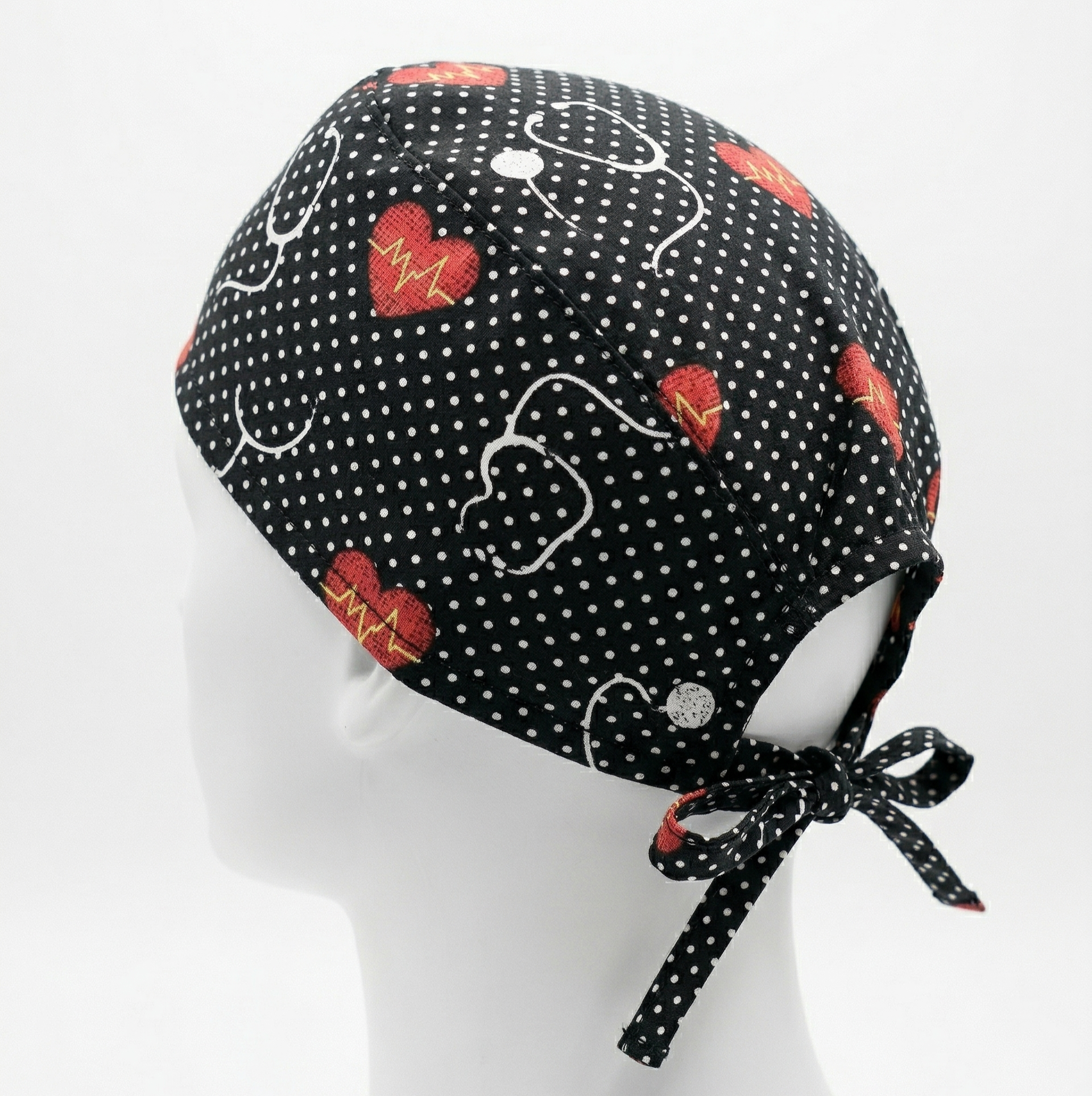 Black Stethoscope Surgical Cap 1: A studio photograph featuring a black surgical cap with a pattern of white stethoscopes and red hearts, displayed on a white mannequin head against a light grey background. The back ties of the cap are clearly visible.