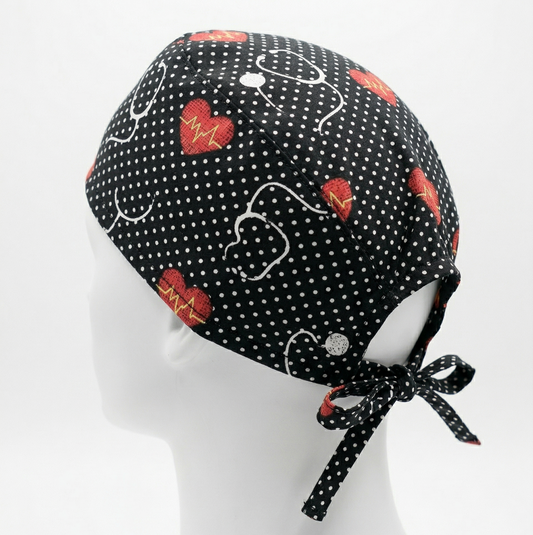 Black Stethoscope Surgical Cap 1: A studio photograph featuring a black surgical cap with a pattern of white stethoscopes and red hearts, displayed on a white mannequin head against a light grey background. The back ties of the cap are clearly visible.