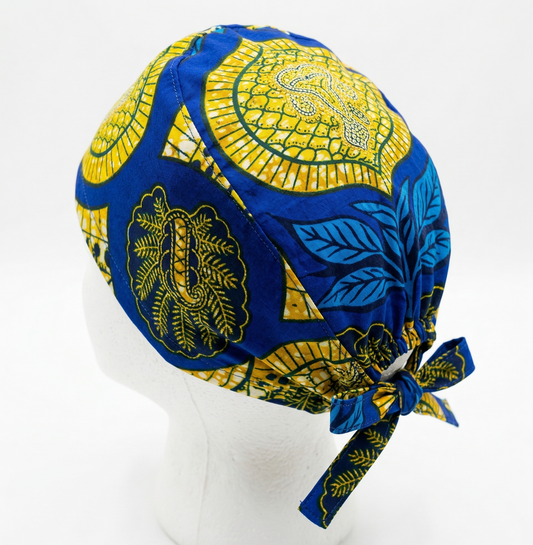 Blue & Yellow African Print Surgical Cap 1: A studio product shot of a surgical cap featuring a vibrant blue, yellow, and red African-inspired pattern. It is displayed on a white mannequin head against a clean white background, showing the tie-back detail.
