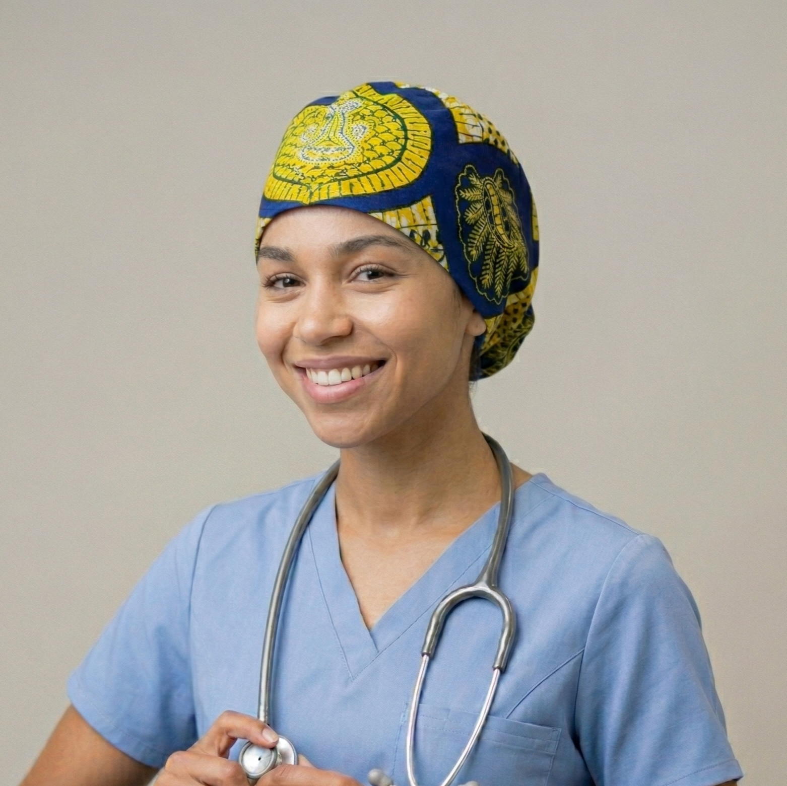 Blue & Yellow African Print Surgical Cap 2: A medium portrait of a smiling female healthcare professional wearing light blue scrubs and a stethoscope. She is wearing the blue and yellow patterned surgical cap, posed against a neutral grey studio background.