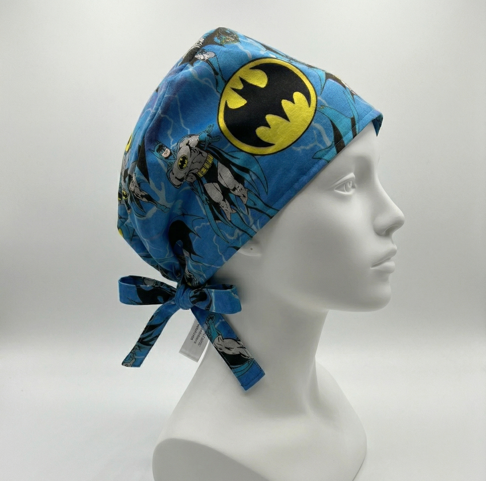 Side profile of a blue Batman themed surgical cap on a white mannequin head, showing the classic yellow bat logo and adjustable tie-back closure.