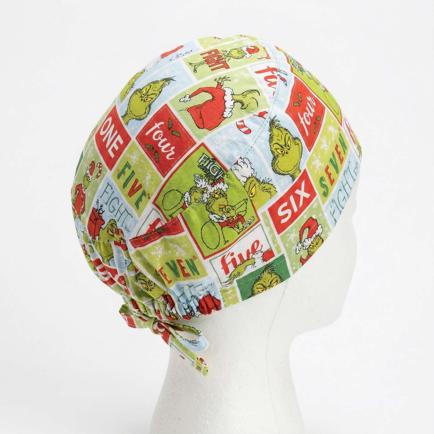 Side profile view of a surgical cap featuring a green Grinch cartoon design on a white mannequin head with adjustable tie-backs.