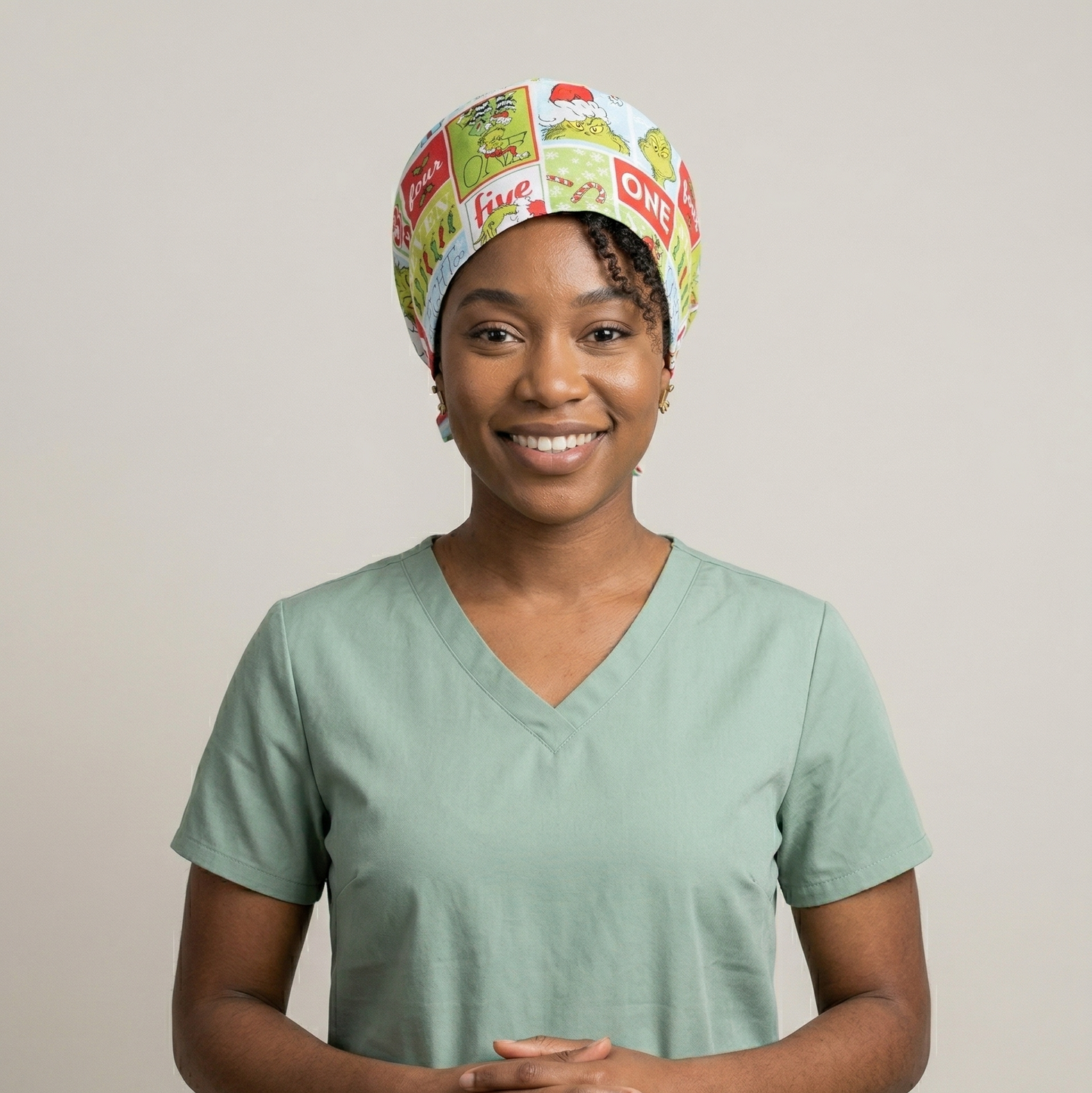 A female model wearing the green Grinch cartoon surgical cap and blue medical scrubs against a neutral studio background.