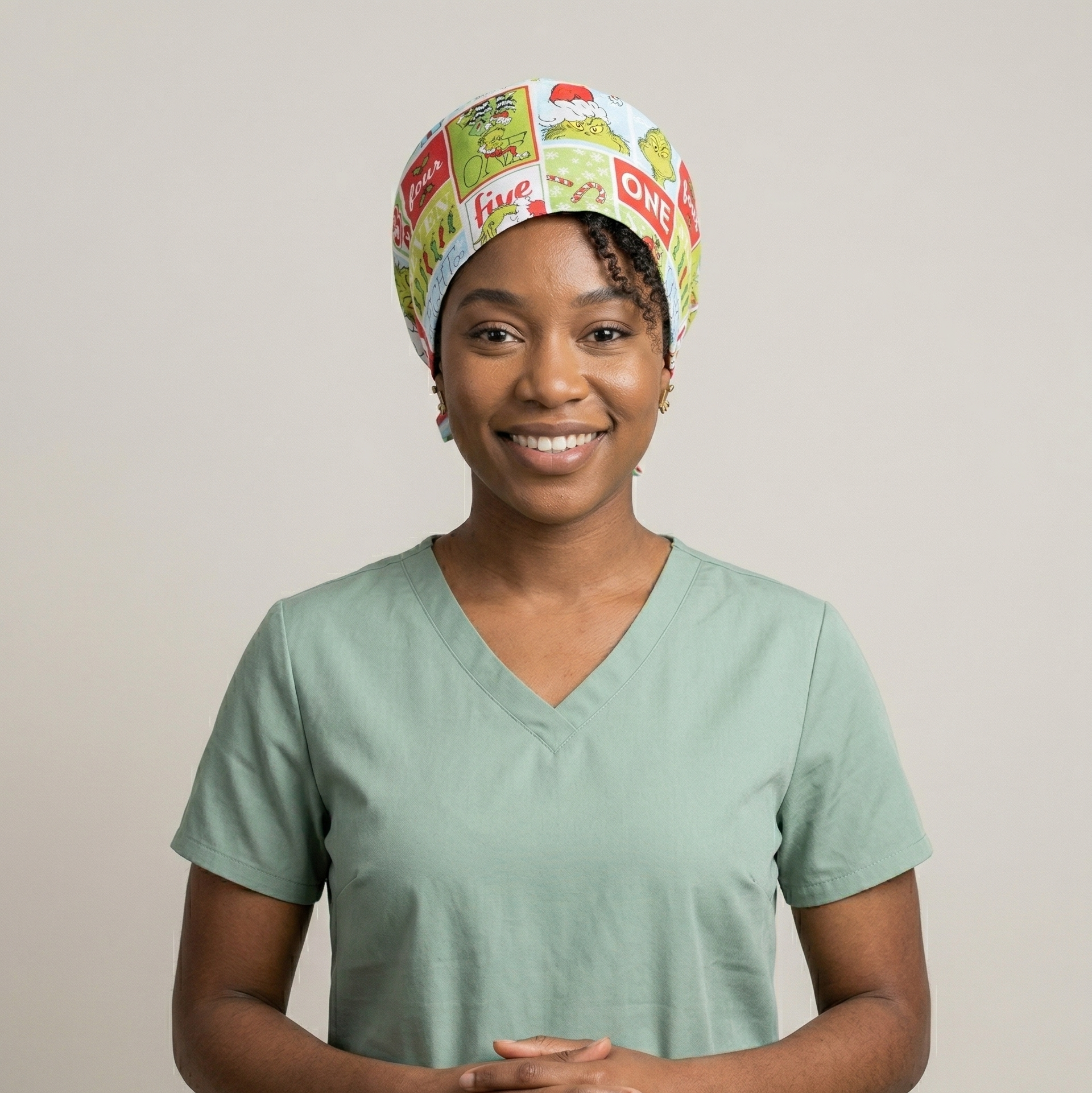 A female model wearing the green Grinch cartoon surgical cap and blue medical scrubs against a neutral studio background.